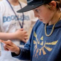 indoor photo at eye-level with a 6th grade student interacting with a touch-screen on a microscope, while another student talks behind him